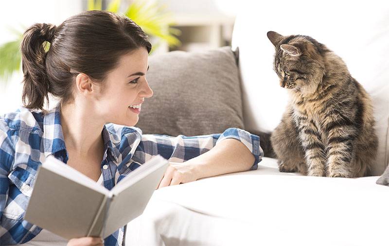 Woman seated on floor holds an open book while smiling at striped cat sitting on a couch.