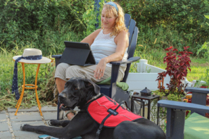 Woman with a laptop sitting outside with her black lab service dog lying at her feet wearing a red harness